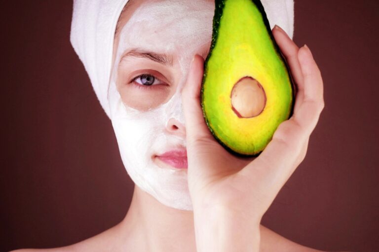 Woman applying a cleansing face mask while holding a cut avocado, symbolizing natural skincare and health.